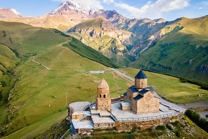 Zu erkennen ist eine historische Kirche mit freistehendem Glockenturm auf einem Hochplateau inmitten einer weiten Berglandschaft. Im Hintergrund erheben sich grüne Berghänge und ein markanter, teilweise schneebedeckter Gipfel unter einem leicht bewölkten Himmel. ausgefallene reiseziele georgien