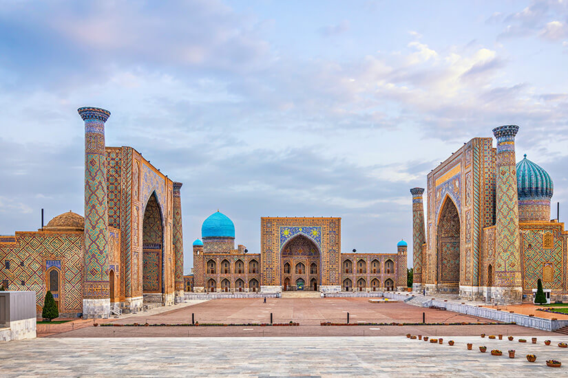 Zu erkennen ist ein weitläufiger historischer Platz, umgeben von prächtigen islamischen Bauwerken mit hohen Portalen, Minaretten und türkisfarbenen Kuppeln. Die Fassaden sind reich mit farbenfrohen geometrischen Mosaiken und Ornamenten verziert. interessante reiseziele usbekistan