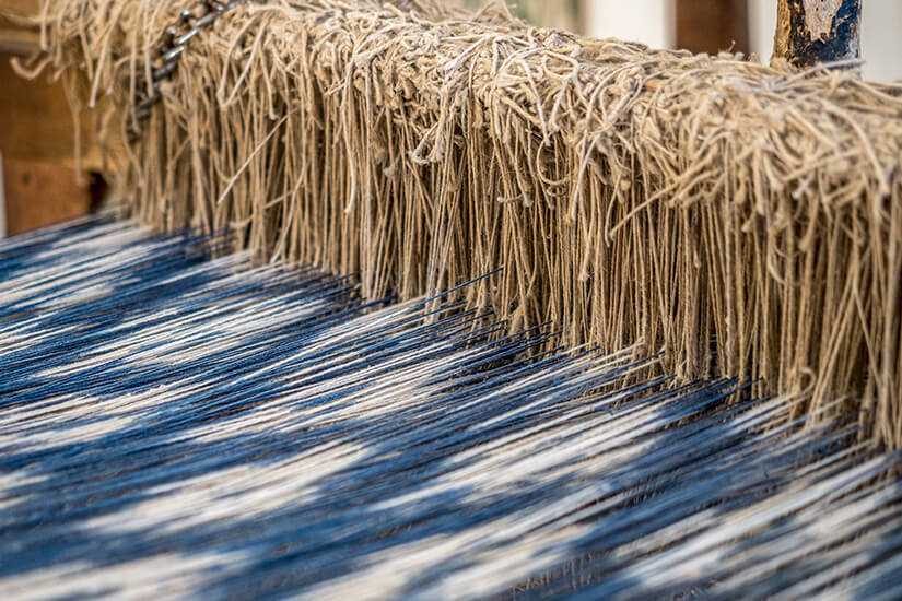 Über einen hölzernen Kettbaum fällt ein dichter Vorhang naturfarbener Hanfschnüre, während darunter die aufgespannte Kette in blau-weißen Zickzackstreifen verläuft. Mallorca Souvenirs Ikat