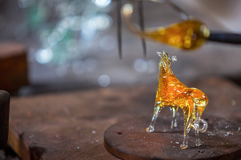 Auf einer runden Holzunterlage steht eine noch glühende Glasfigur in Pferdegestalt, deren bernsteinfarbener Körper von innen zu leuchten scheint. Mallorca Souvenirs Gordiola