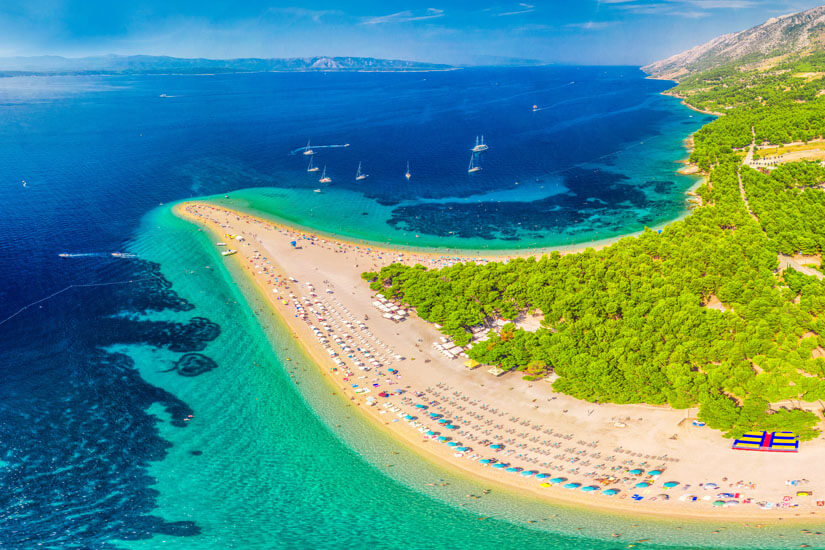 Bunte Sonnenschirme und Liegen säumen beide Seiten des Strands, dahinter erstreckt sich dichter Pinienwald, und weiter draußen ankern Segelboote vor der bergigen Küste am Horizont. Reiseziele September Kroatien