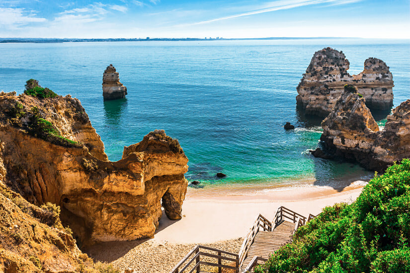 Zerklüftete goldgelbe Sandsteinfelsen mit Torbögen und freistehenden Felsnadeln rahmen eine kleine Sandbucht mit türkisgrünem Wasser. Reiseziele September Algarve