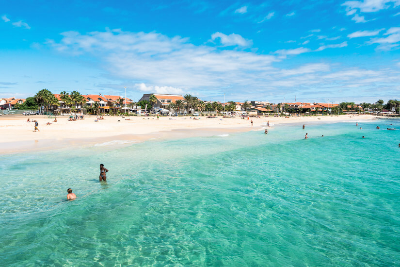 Türkisgrünes, kristallklares Flachwasser reicht bis an einen weiten hellen Sandstrand, in dem einzelne Badende stehen und schwimmen, während weiter hinten Menschen zwischen Sonnenschirmen und Liegen verteilt sind. Reiseziele September Kapverden