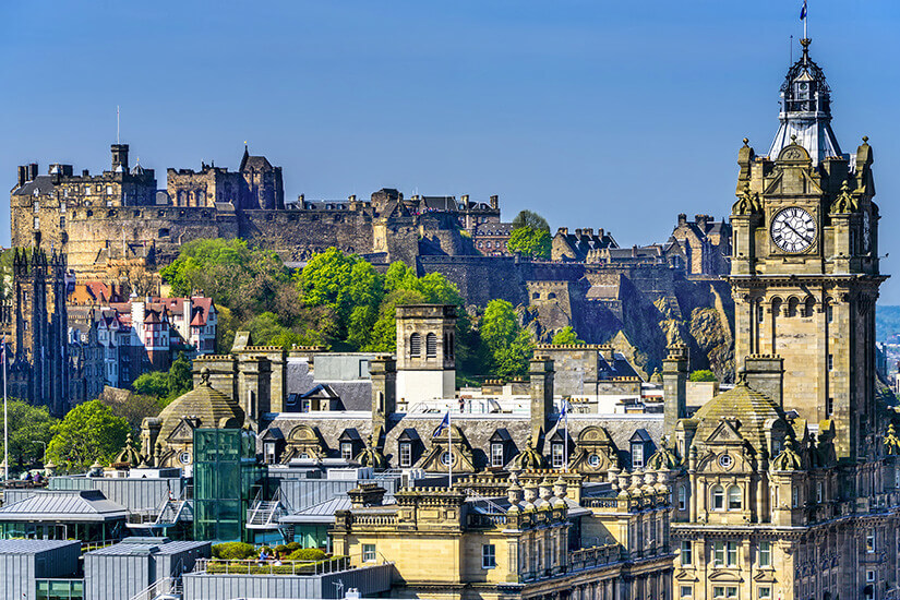 Über der Dächerlandschaft Edinburghs thront auf steilem Vulkanfelsen die massive Burganlage, während rechts der markante Uhrturm des Balmoral Hotels mit seinen Türmchen und der Fahne emporragt. Reiseziele September Edinburgh