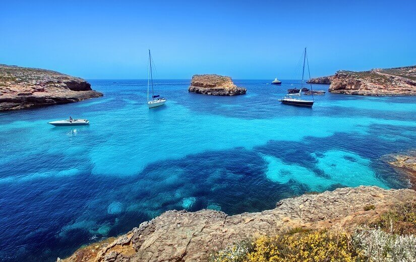 Leuchtend türkisfarbenes, glasklares Wasser füllt eine flache Bucht, in der zwei Segelboote und ein Motorboot vor Anker liegen. Herbstferien Malta