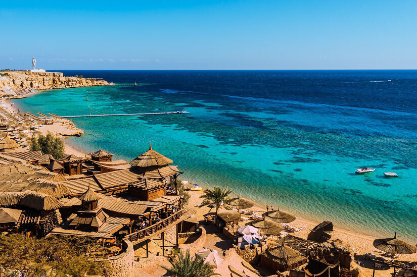 Von einer Felsterrasse öffnet sich der Blick über eine Bucht am Roten Meer, deren Wasser vom seichten Türkis über dunkle Korallenflecken bis ins tiefe Blau übergeht. Herbstferien Aegypten