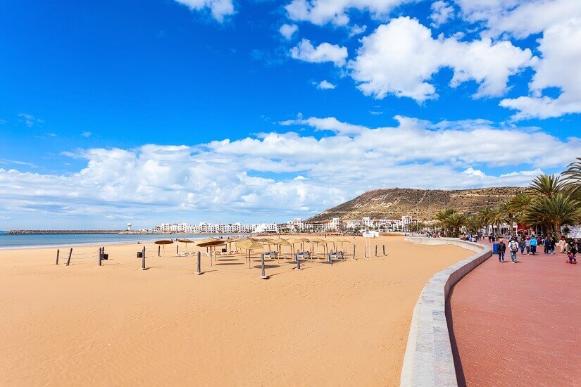 Weiter goldgelber Sandstrand mit vereinzelten Strohschirmen zieht sich in sanftem Bogen zur Bucht, während rechts Spaziergänger auf einer rotbraunen, palmengesäumten Promenade entlanggehen. Herbstferien Agadir