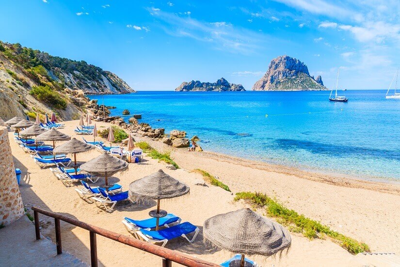 Blaue Liegen unter geflochtenen Strohschirmen reihen sich am hellen Sand der Cala d'Hort auf Ibiza, eingefasst von einem Holzgeländer und felsigen Hängen mit Pinien. Herbstferien Ibiza
