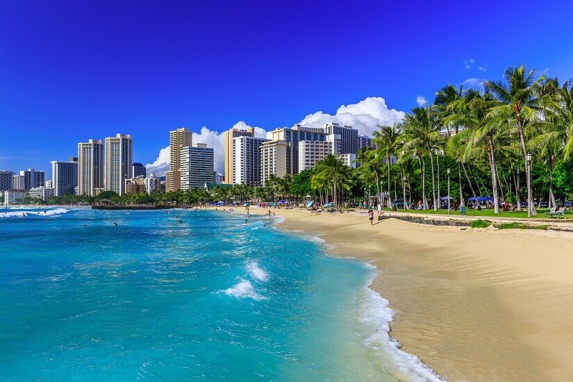 Türkisblaue Wellen rollen schäumend auf den breiten goldenen Sand des Waikiki Beach, an dem einzelne Badegäste und Spaziergänger unterwegs sind. Herbstferien Hawaii