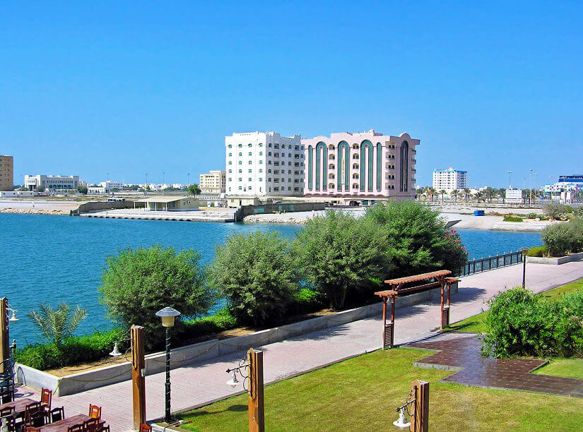 Entlang eines türkisgrünen Meeresarms zieht sich eine gepflasterte Uferpromenade mit Rasenflächen, Laternen, einer hölzernen Pergola und niedrigen Sträuchern. Herbstferien VAE