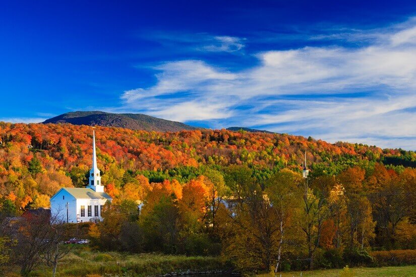 Vor bewaldeten Hügeln in leuchtendem Herbstlaub aus Rot, Orange und Gelb steht eine weiße Holzkirche mit spitzem Turm. Eine klassische Neuengland-Szenerie, wie man sie aus Stowe in Vermont kennt. Herbstferien USA