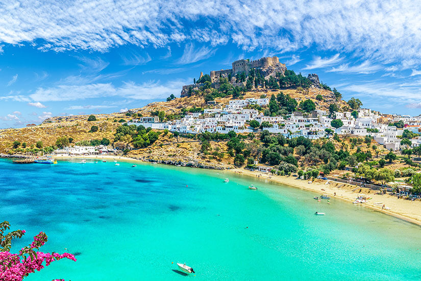 Unterhalb der Akropolis von Lindos leuchten weiße Häuser an einem sonnenverwöhnten Hang, während davor eine geschützte Bucht in Türkis schimmert. Das Panorama vereint antike Geschichte, typisch griechische Architektur und mediterrane Küstenlandschaft. Griechsiche Inseln Rhodos