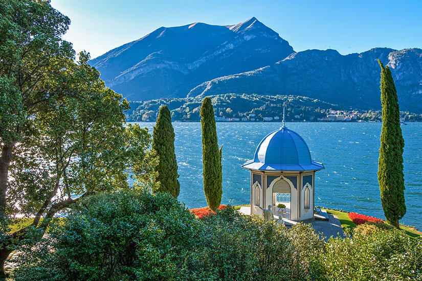 Ein eleganter Pavillon mit blauem Kuppeldach steht direkt am Ufer des Comer Sees in Bellagio. Schlanke Zypressen und gepflegte Blumenbeete umgeben das kleine Bauwerk, während im Hintergrund das tiefblaue Wasser und eine imposante Bergkulisse unter strahlendem Himmel zu sehen sind. Bellagio Italien Willkommen