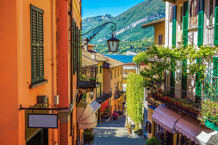Blick steil hinab durch eine malerische, schmale Treppengasse in der Altstadt von Bellagio. Die bunten Häuserfronten in warmen Terracotta-, Gelb- und Weißtönen sind mit grünen Klappläden, geschmiedeten Balkonen und üppig blühenden Pflanzen verziert. Am Ende der Gasse gibt der Blick den Comer See und die gegenüberliegende Bergkulisse frei. Bellagio Italien Altstadt