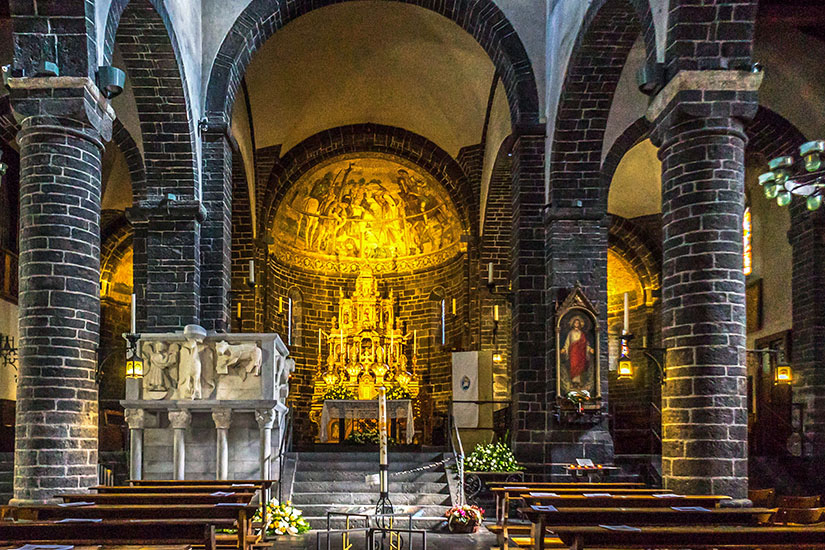 Der stimmungsvoll ausgeleuchtete Innenraum der romanischen Basilica San Giacomo in Bellagio. Mächtige Säulen und Rundbögen aus dunklem Naturstein gliedern das Schiff. Im Zentrum strahlt der vergoldete Hauptaltar unter einer prächtigen, goldglänzenden Apsismalerei, während im Vordergrund hölzerne Kirchenbänke stehen. Bellagio Italien Basilica San Giacomo