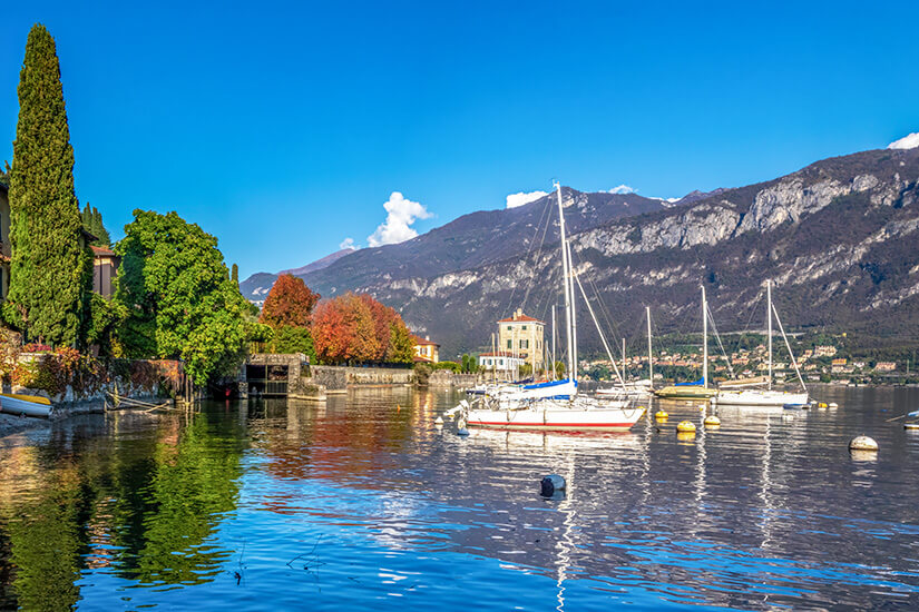 Sonniges Seeufer im malerischen Fischerdorf Pescallo nahe Bellagio am Comer See. Mehrere Segelboote ankern im ruhigen, tiefblauen Wasser. Das begrünte Ufer mit hohen Zypressen und herbstlich gefärbten Bäumen spiegelt sich auf der Wasseroberfläche, während im Hintergrund imposante Felsberge aufragen. Bellagio Italien Boote