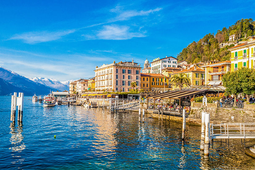 Panoramablick auf die malerische Uferfront von Bellagio am Comer See. Elegante, pastellfarbene Gebäude und Terrassencafés reihen sich direkt am Wasser aneinander, während Ausflugsschiffe am Anleger anlegen. Im Hintergrund erheben sich bewaldete Hügel und schneebedeckte Berggipfel unter einem strahlend blauen Himmel. Bellagio Italien Seeblick
