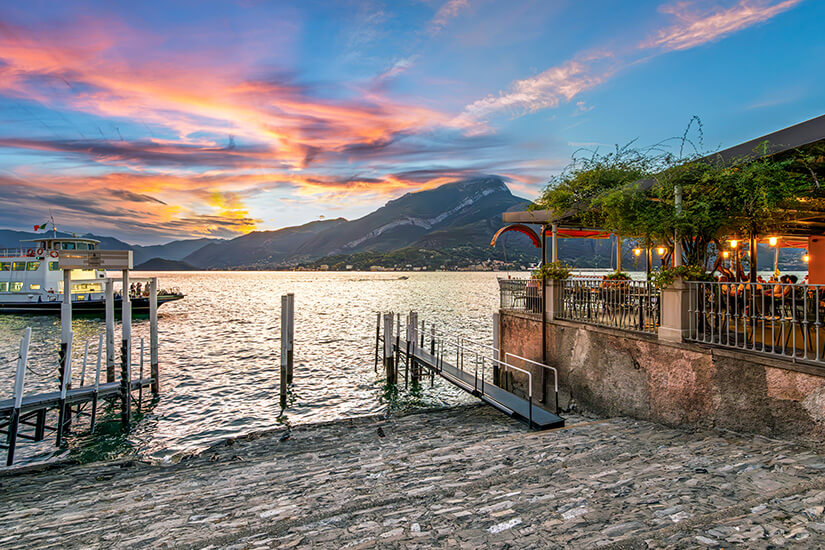 Ein Restaurant am Ufer des Comer Sees in Bellagio während eines stimmungsvollen Sonnenuntergangs. Der Himmel leuchtet in rosa und orangefarbenen Tönen über den Silhouetten der Berge. Rechts sitzen Gäste auf der beleuchteten Terrasse direkt am Wasser, während links eine Fähre am Steg im sanft glitzernden See liegt. Bellagio Italien Restaurant