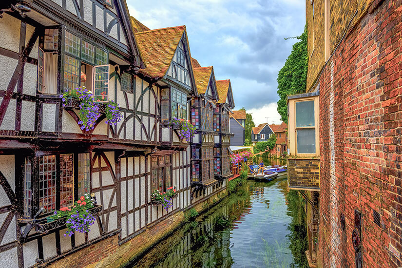 Zu erkennen ist eine schmale Wasserstraße, die von historischen Fachwerkhäusern mit Blumenschmuck und alten Backsteinmauern gesäumt wird. Im Hintergrund liegen kleine Boote auf dem Wasser, während sich weitere malerische Häuser entlang des Kanals erstrecken. Canterbury Weberhaeuser