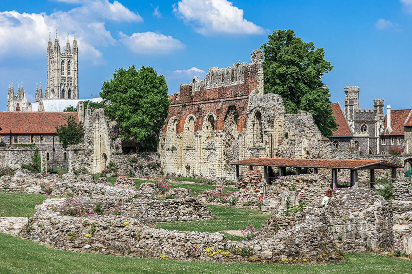 Zu erkennen sind die Ruinen einer historischen Klosteranlage mit alten Steinmauern, Bögen und Fundamentresten, zwischen denen Blumen und Gras wachsen. Im Hintergrund erhebt sich der markante Turm einer großen gotischen Kathedrale. Canterbury Kloster