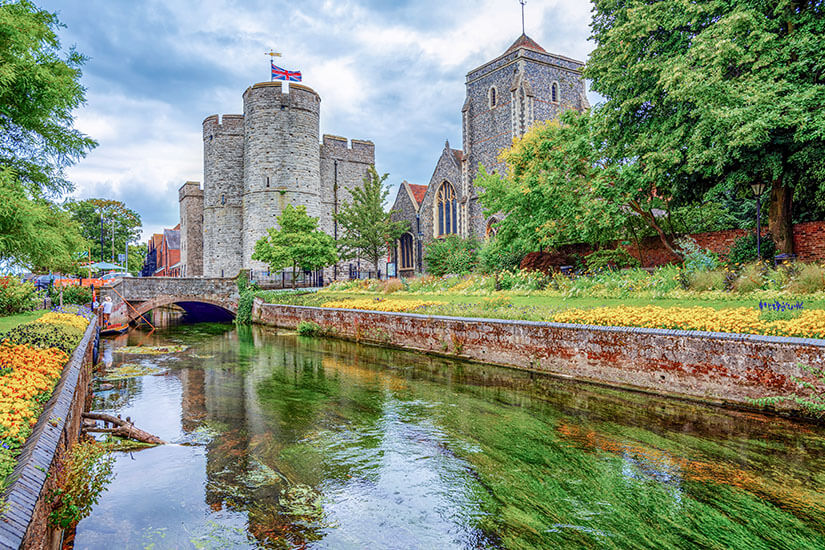 Zu erkennen sind ein historisches Stadttor mit Rundtürmen und eine alte Kirche an einem schmalen Fluss. Üppige Bäume, Blumenbeete und eine kleine Brücke prägen die idyllische Umgebung. Canterbury Westgate