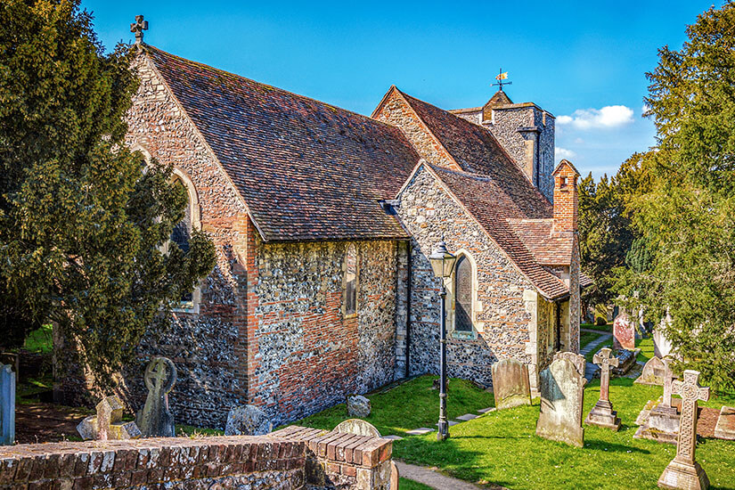 Zu erkennen ist eine historische Steinkirche mit roten Ziegeldächern, umgeben von einem alten Friedhof. Zahlreiche verwitterte Grabsteine und Kreuze stehen zwischen Bäumen und gepflegten Grünflächen. Canterbury Kirche