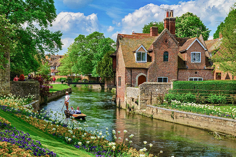 Zu erkennen ist ein idyllischer Flusslauf, gesäumt von historischen Backsteinhäusern, gepflegten Gärten und bunten Blumenbeeten. Auf dem Wasser fährt ein kleines Stocherboot mit mehreren Personen. Kent Punting