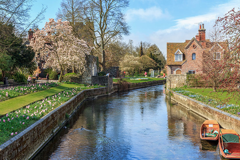 Zu erkennen ist ein idyllischer Flusslauf, gesäumt von blühenden Gärten, alten Bäumen und historischen Backsteinhäusern. Am Ufer liegen kleine Holzboote, während Frühlingsblumen und blühende Bäume die Szenerie prägen. Kent Fruehling