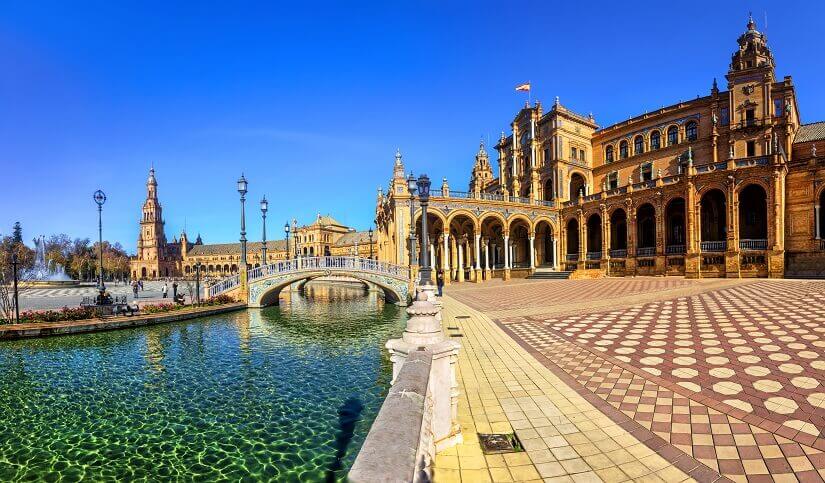 Der halbrunde Wasserkanal der Plaza de España in Sevilla zieht sich unter einer gekachelten Brücke hindurch, gesäumt von gemustertem Pflaster und schmiedeeisernen Laternen. Herbstferien Sevilla