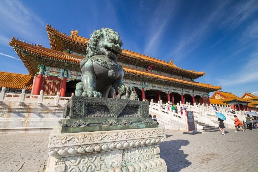 Auf einem reich verzierten Marmorsockel wacht ein bronzener Wächterlöwe vor einer kaiserlichen Halle der Verbotenen Stadt in Peking, deren geschwungene Dächer mit goldgelben Ziegeln und bunt bemalten Balken über roten Säulen ruhen. Herbstferien Peking
