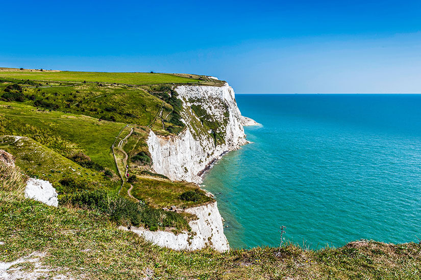 Zu erkennen sind markante weiße Kreidefelsen, die steil zum türkisblauen Meer abfallen. Über die grünen Klippen führt ein schmaler Wanderweg entlang der Küste. England Dover