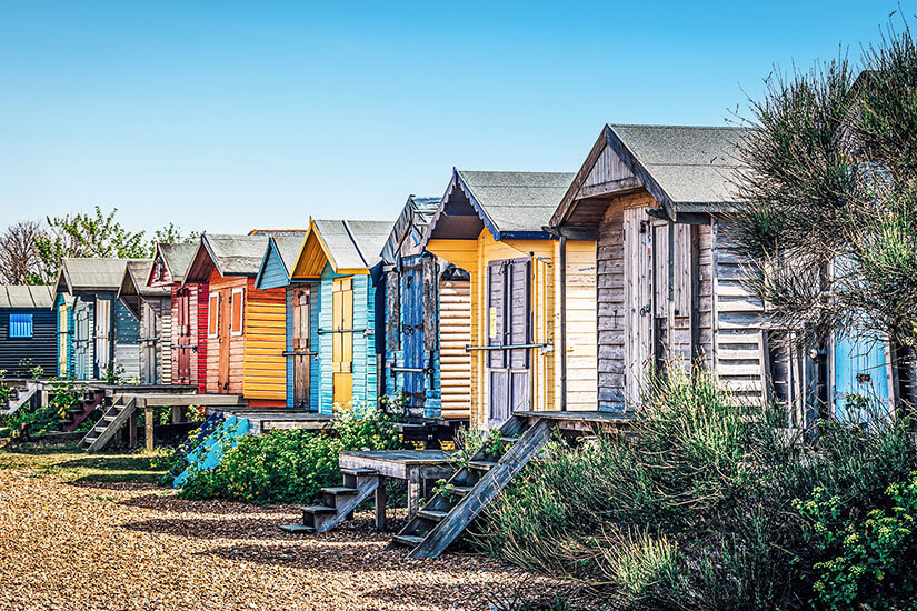 Zu erkennen ist eine Reihe kleiner, farbenfroher Strandhütten aus Holz mit spitzen Dächern. Die Hütten stehen dicht nebeneinander an einem kiesigen Küstenabschnitt und sind über kleine Treppen zugänglich. England Whitstable