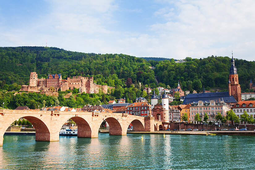 Die Stadt Heidelberg vom Flussufer aus betrachtet. Das Wasser leuchtet grünblau. Die Brücke spiegelt sich im Fluss und das Schloss thront im Grünen. Schoenste Staedte Deutschlands Heidelberg