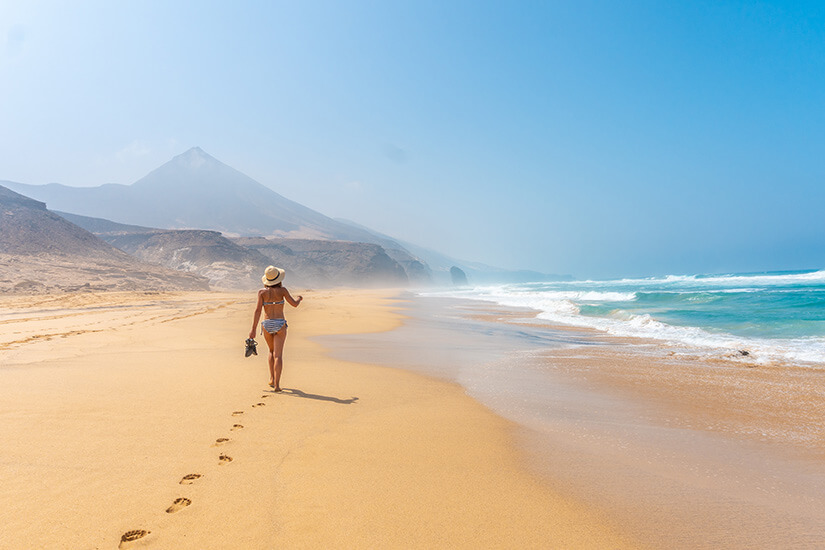 Eine Frau im Bikini mit Strohhut und Sandalen in der Hand geht barfuß über einen menschenleeren goldgelben Sandstrand und hinterlässt eine Spur von Fußstapfen. Wo ist es im Winter warm Fuerteventura