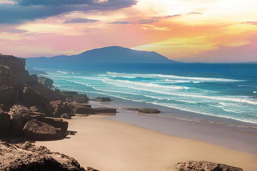 In warmen Rosa-, Orange- und Goldtönen glüht der Abendhimmel über einer einsamen Atlantikbucht, in der türkisfarbene Brandungswellen in mehreren Reihen auf den hellen Sand rollen. Wo ist es im Winter warm Agadir