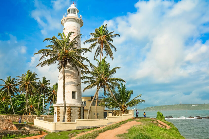 Zwischen hohen Kokospalmen ragt der weiße Leuchtturm von Galle auf der historischen Festungsmauer empor, umgeben von einem gelben Nebengebäude und grünen Grasflächen. Wo ist es im Winter warm Sri Lanka