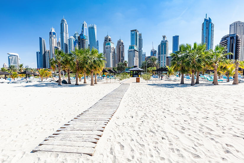 Über weißen Sand führt ein hölzerner Bohlenweg schnurgerade auf eine Reihe Palmen zu, hinter denen Liegen, Sonnenschirme und ein Pavillon einer Strandanlage stehen. Wo ist es im Winter warm Dubai