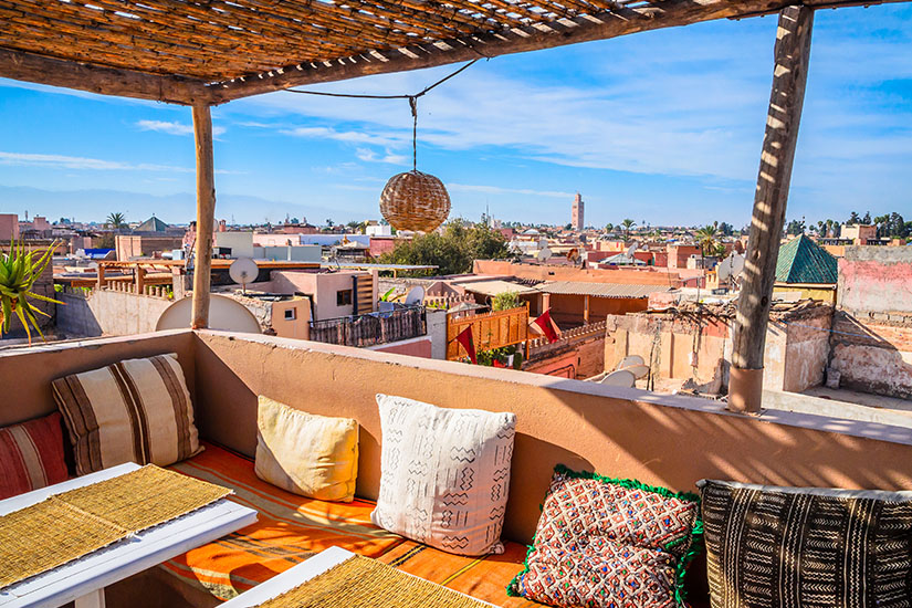Von einer schattigen Dachterrasse mit Schilfpergola, Rattanlampe und bunten Sitzkissen auf lehmfarbenen Bänken schweift der Blick über die Dächer der Medina von Marrakesch. Wo ist es im Winter warm Marrakesch