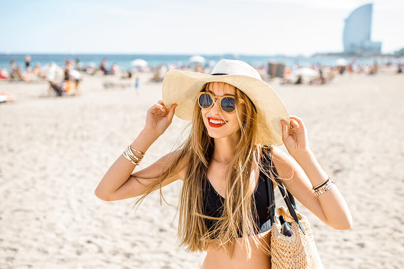 Lächelnd hält eine junge Frau mit langen blonden Haaren, runder Sonnenbrille und schwarzem Bikini ihren breitkrempigen Strohhut fest, über der Schulter hängt eine geflochtene Strandtasche. Singlereisen Barcelona