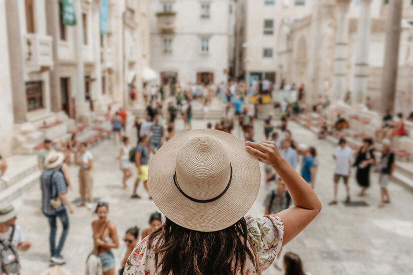 Von hinten fotografiert hält eine dunkelhaarige Frau im geblümten Sommerkleid ihren breitkrempigen Strohhut fest und blickt auf das Peristyl im Diokletianpalast von Split hinab. Singlereisen Split