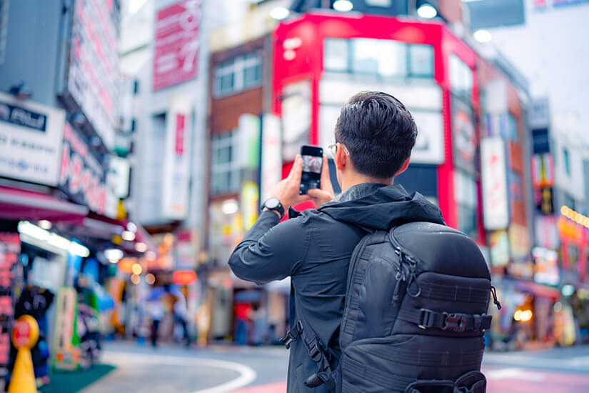 Von hinten aufgenommen fotografiert ein junger Mann mit schwarzer Jacke und großem Rucksack mit seinem Smartphone die Häuserfront einer belebten japanischen Einkaufsstraße. Singlereisen Tokio