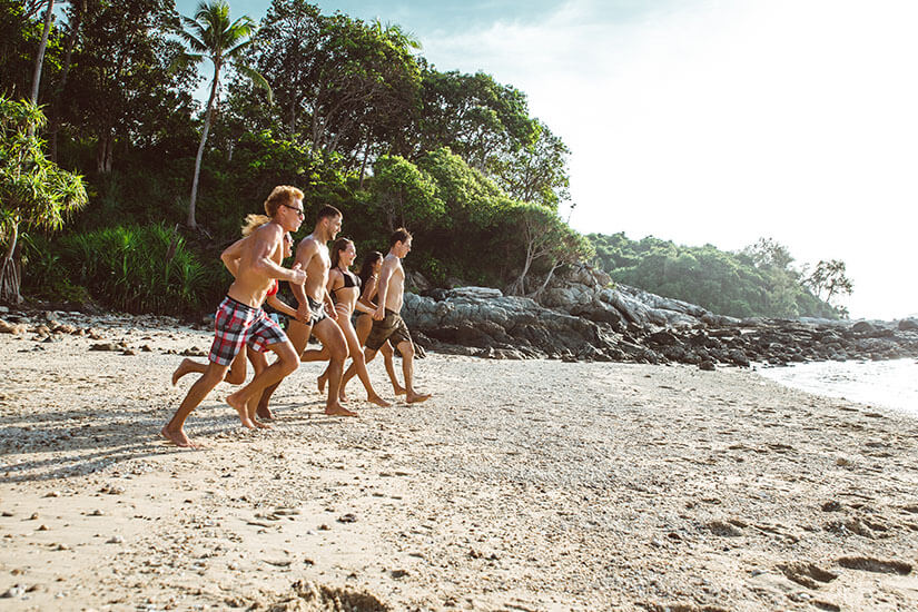 Fünf junge Leute in Badebekleidung laufen barfuß hintereinander über einen hellen Sandstrand, offenbar auf dem Weg ins Wasser. Singlereisen Bali