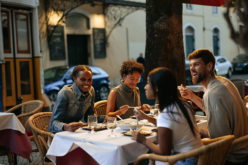 Vier junge Leute sitzen lachend und plaudernd in Korbstühlen an einem weiß gedeckten Tisch vor einem Restaurant und teilen sich mehrere Gerichte samt Wein und Wasser. Singlereisen Lissabon
