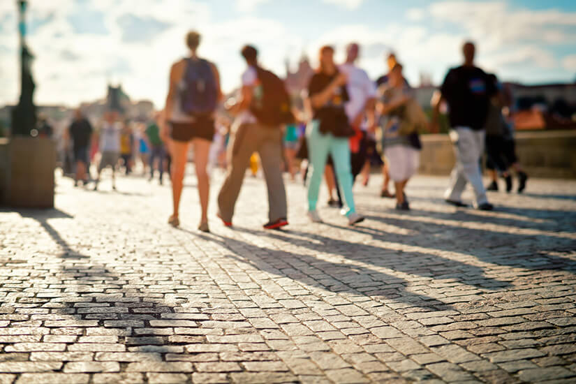 Scharf gestochenes Kopfsteinpflaster füllt den Vordergrund, darüber bewegen sich unscharfe Passanten in sommerlicher Kleidung im Gegenlicht über eine belebte Brücke. Singlereisen Prag