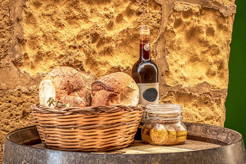 Zu erkennen sind rustikale Brotlaibe in einem Weidenkorb, eine Flasche Wein und ein Glas mit eingelegten Lebensmitteln. Die Anordnung vor einer groben Steinwand vermittelt eine mediterrane, traditionelle Atmosphäre. Malta Essen Traditionen
