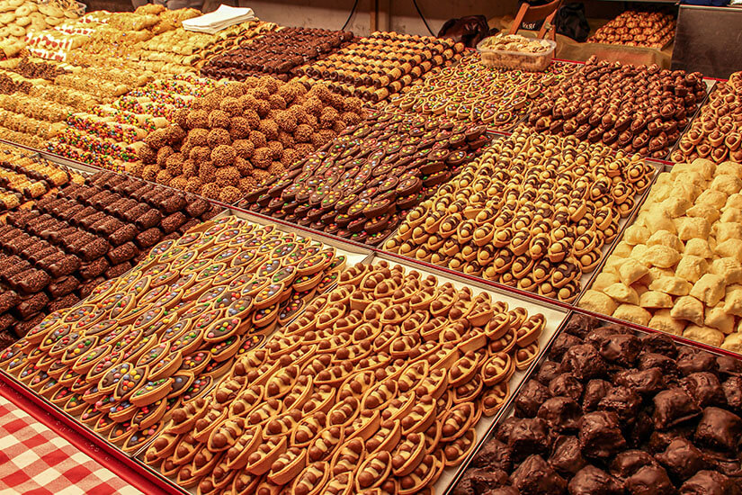 Zu erkennen ist eine große Auswahl an Gebäck und Süßwaren, darunter Kekse, schokolierte Stücke und bunt verzierte Plätzchen. Die verschiedenen Sorten sind dicht nebeneinander auf einem Marktstand ausgelegt. Malta Essen Spezialitaeten