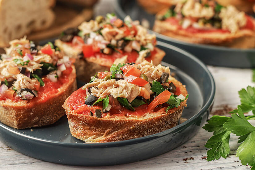 Zu erkennen sind geröstete Brotscheiben mit Tomatenaufstrich, belegt mit Thunfisch, Tomaten, Oliven, Zwiebeln und frischen Kräutern. Im Hintergrund liegen weitere belegte Brote auf Tellern. Malta Essen Typisch Ftira