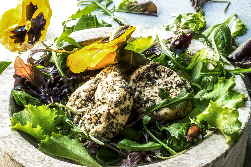 Zu erkennen sind mehrere mit Kräutern und Pfeffer gewürzte Käsetaler auf einem frischen Blattsalat. Essbare gelbe Blüten und verschiedene Salatblätter sorgen für eine farbenfrohe Garnierung. Maltesisch Essen Gbejna