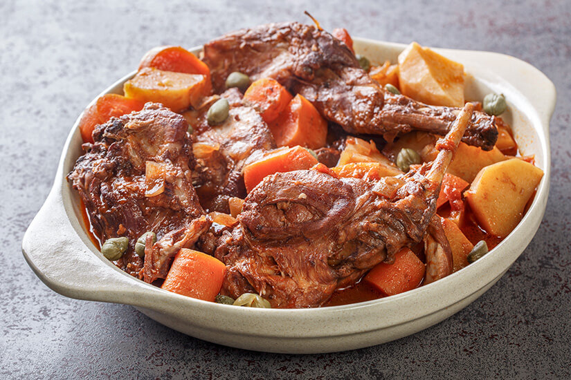 Zu erkennen ist ein geschmortes Fleischgericht mit mehreren Fleischstücken, Kartoffeln, Karotten und Kapern in einer kräftigen Tomatensoße. Es wird in einer großen, rustikalen Auflaufform serviert. Essen auf Malta Fenkata