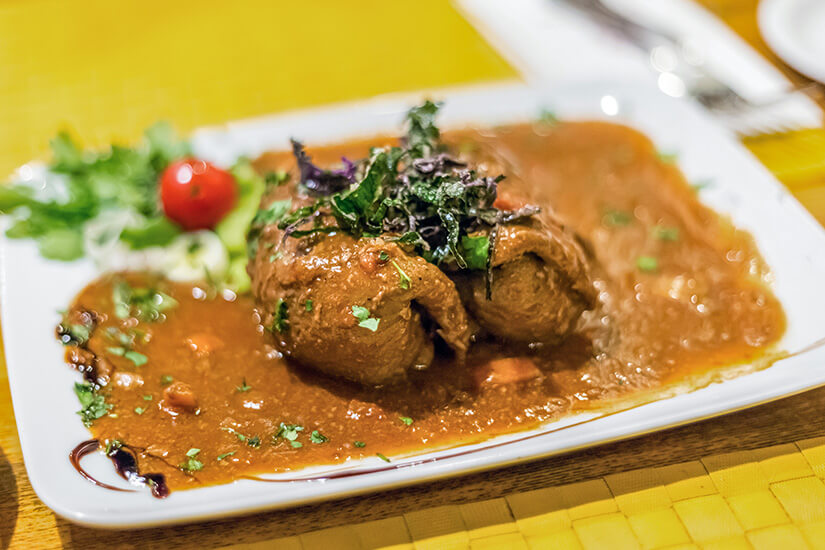 Zu erkennen sind mehrere geschmorte Fleischrouladen in einer kräftigen braunen Soße, garniert mit frischen Kräutern. Dazu werden etwas Salat und eine kleine Tomate serviert. Malta Essen Bragioli